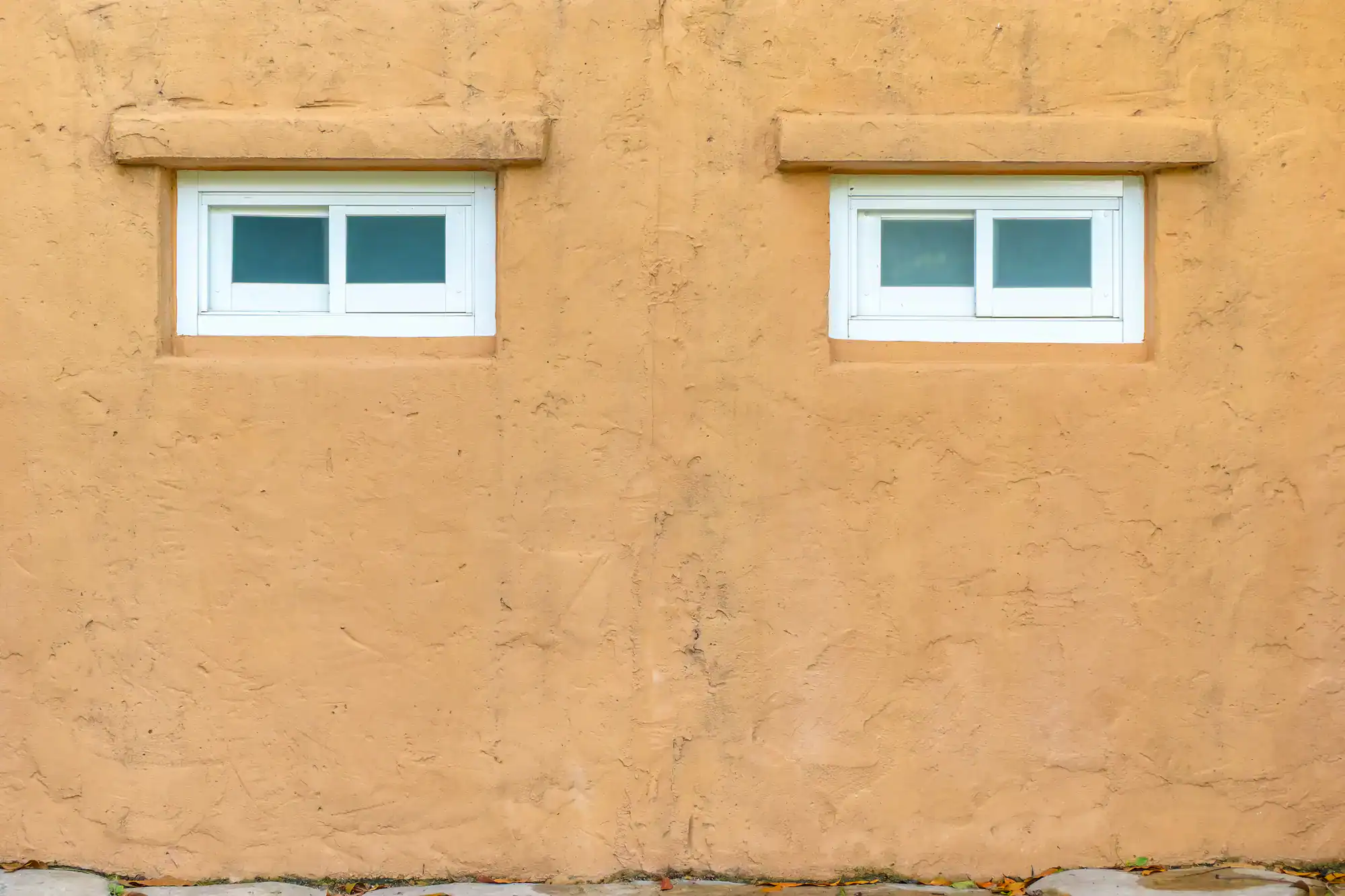Two small rectangular white-framed windows are set into an adobe-style beige stucco wall, positioned side by side with a large blank space between and beneath them, highlighting expert work by a Remodeling Contractor San Diego County, CA.